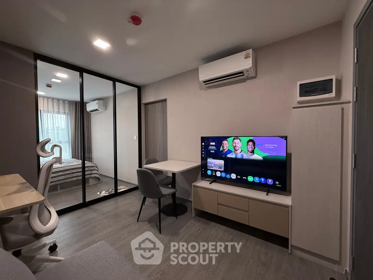 Modern living room with sleek furniture and large TV, adjacent to a cozy bedroom with glass partition.