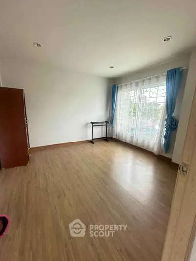 Spacious empty room with wooden flooring and large window with blue curtains.