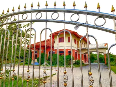 Charming two-story house with red accents and gated entrance, surrounded by lush greenery.