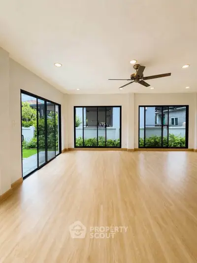 Spacious living room with large windows and garden view, featuring modern ceiling fan and wooden flooring.