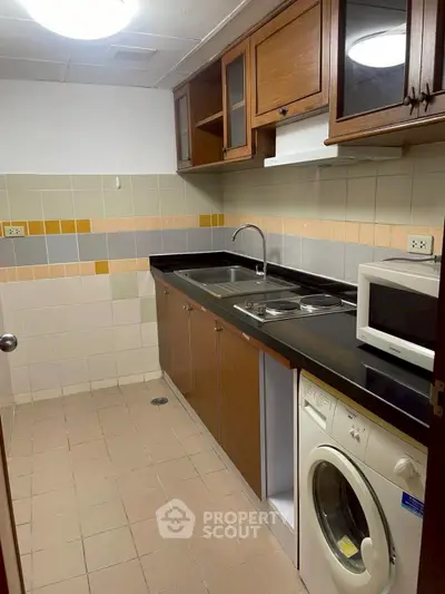 Compact kitchen with wooden cabinets, washing machine, and microwave in a cozy apartment.