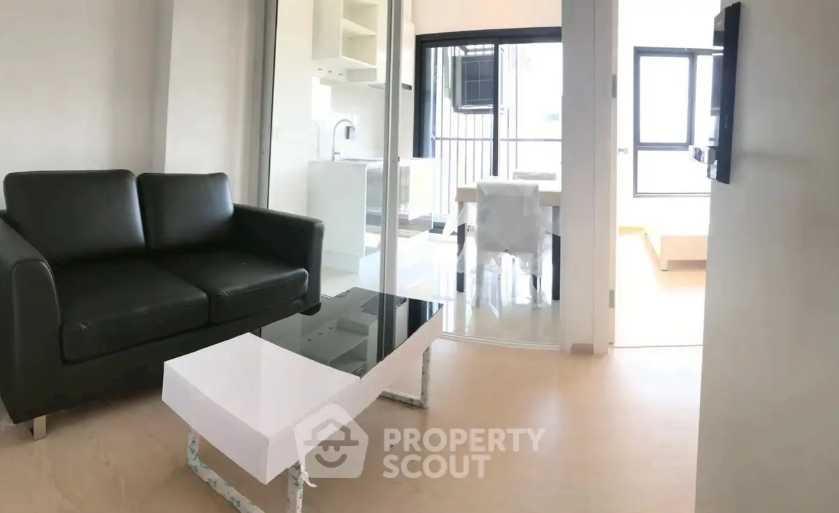 Modern living room with sleek black sofa and glass coffee table, open to bright dining area.