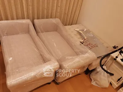 Cozy living room with wrapped furniture ready for setup, featuring elegant beige sofas and a tufted ottoman.