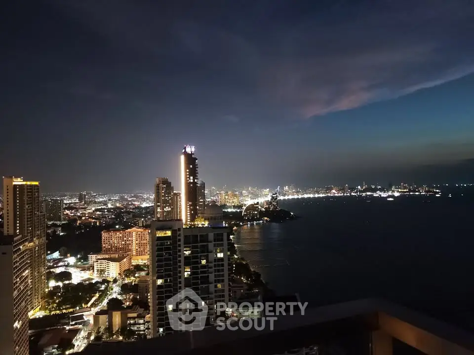 Stunning cityscape view from a high-rise balcony overlooking the ocean at night, showcasing vibrant city lights.