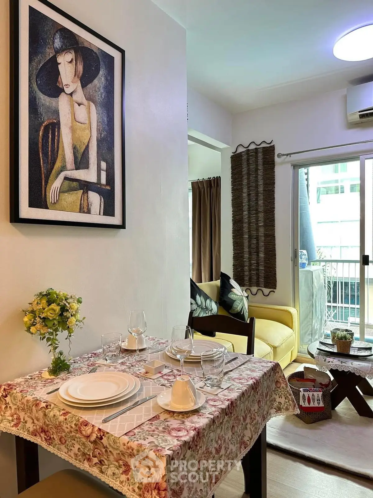Charming dining area with floral tablecloth and art, leading to cozy living room and balcony.