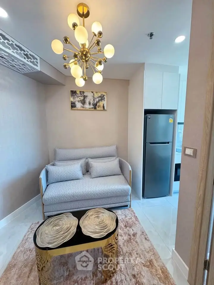 Stylish living room with modern chandelier and cozy sofa, adjacent to a sleek kitchen area with stainless steel fridge.