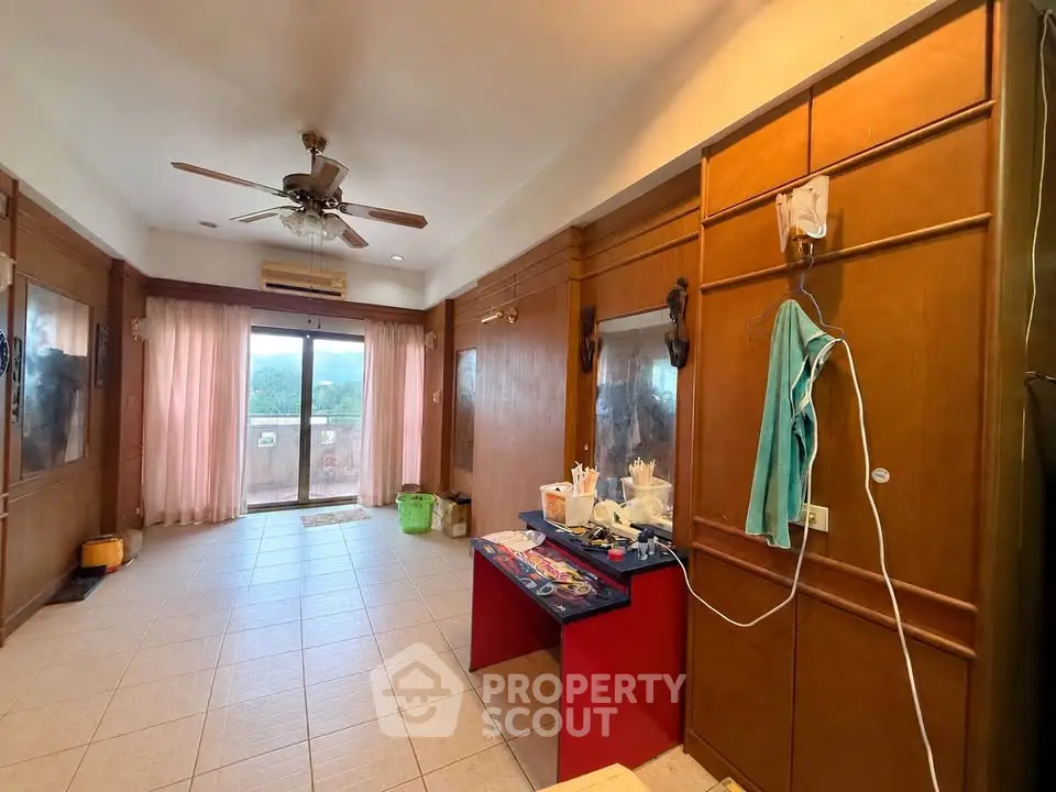Spacious living room with ceiling fan and balcony access, featuring large windows and wooden paneling.