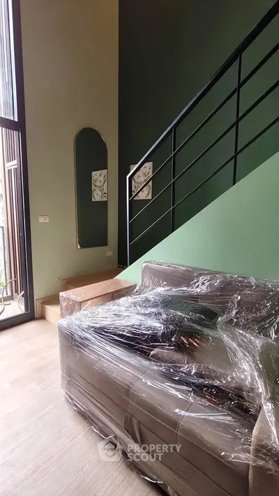 Modern living room with high ceiling and staircase, featuring a wrapped sofa and large window for natural light.