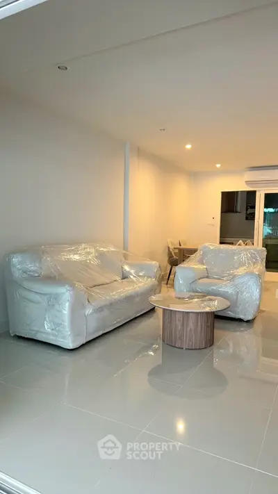 Spacious modern living room with new furniture wrapped in plastic, featuring sleek tile flooring and ambient lighting.