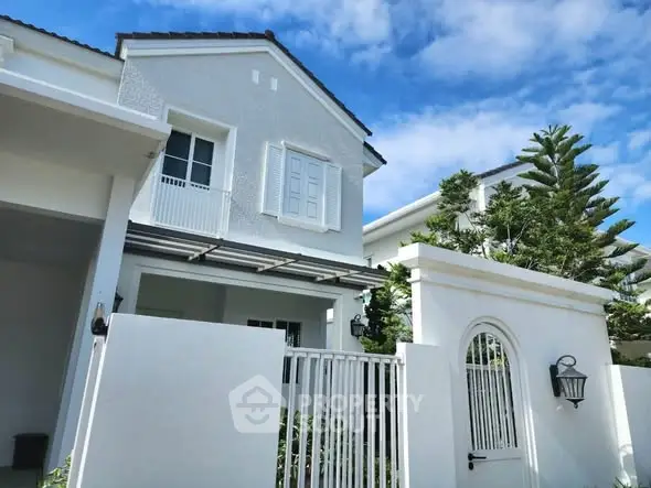 Charming white two-story house with elegant gate and lush greenery under a clear blue sky.