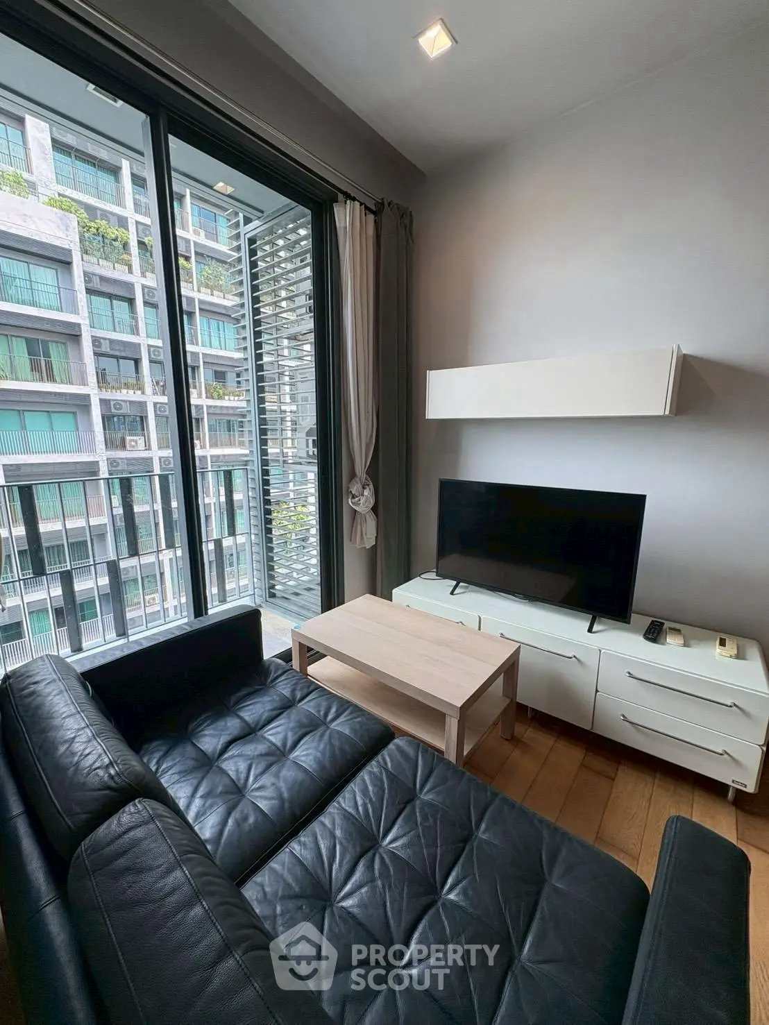 Modern living room with sleek black leather sofa and large TV, featuring floor-to-ceiling windows with city view.