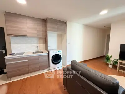 Modern open-layout living room with compact kitchen and washing machine, featuring sleek wooden cabinetry and cozy seating.