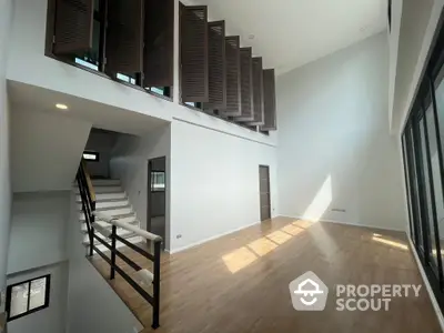 Spacious high-ceiling living room with elegant wooden floors and modern staircase leading to an upper level, bathed in natural light from large windows.