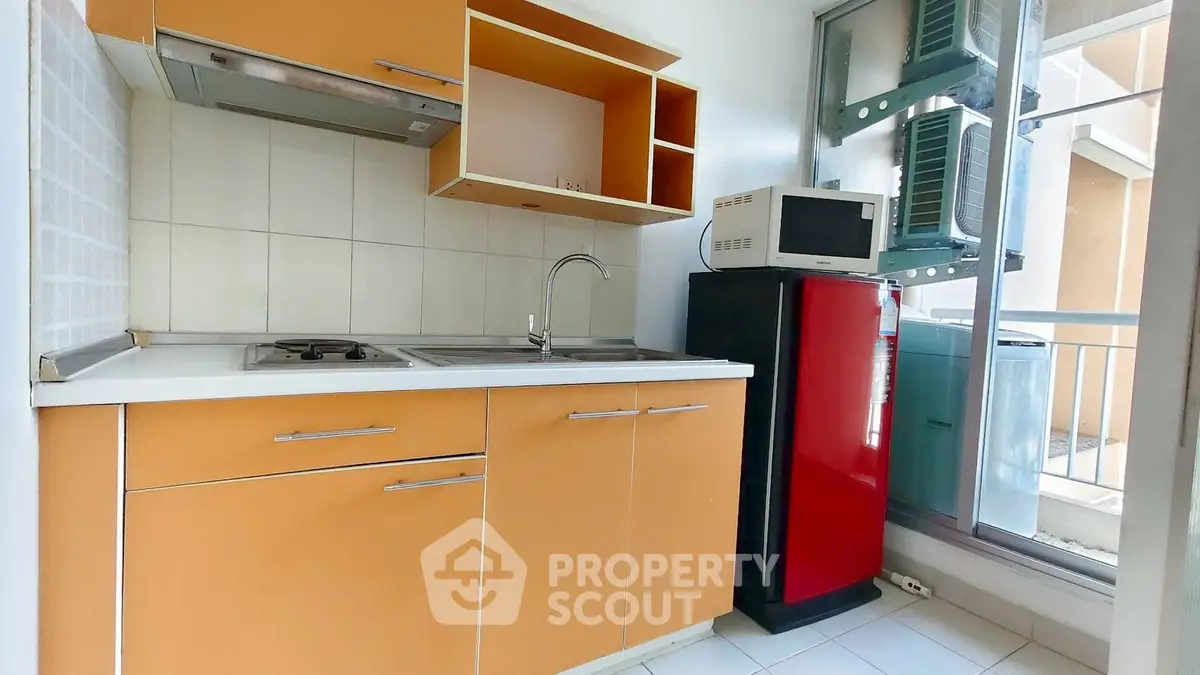 Compact kitchen with orange cabinets, microwave, and red fridge in modern apartment.