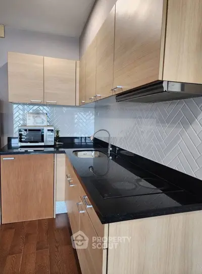 Modern kitchen with sleek black countertops and light wood cabinets, featuring a built-in oven and stylish backsplash.