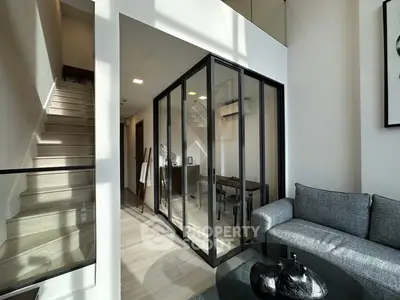 Modern living room with glass-walled study and staircase, featuring sleek furniture and abundant natural light.