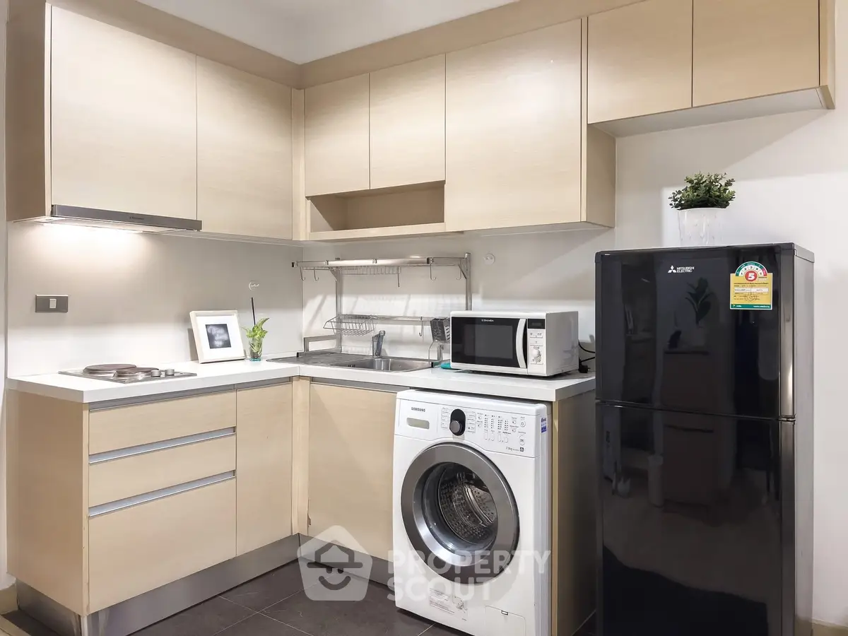 Modern kitchen with light wood cabinets, washing machine, and black fridge in a compact layout.