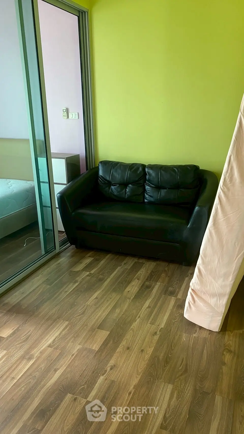 Cozy living room with black leather sofa and wooden flooring, adjacent to a bedroom with glass sliding doors.