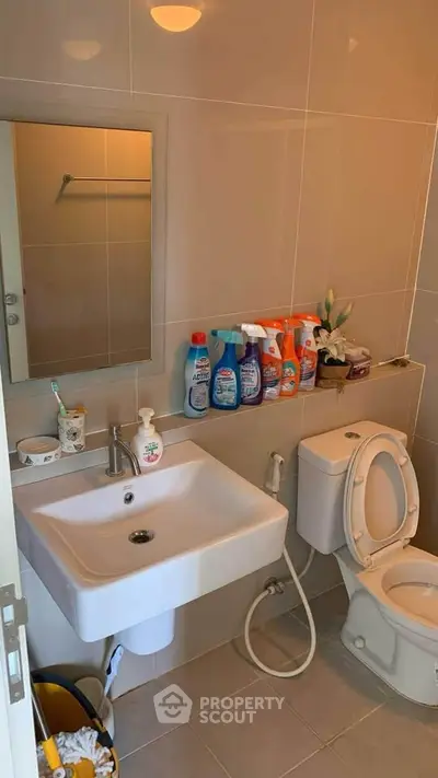 Modern bathroom with sink, toilet, and cleaning supplies on shelf.