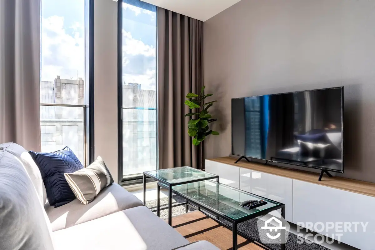 Bright and modern living room with large windows offering ample natural light, sleek furnishings, and a wall-mounted TV, perfect for urban living.