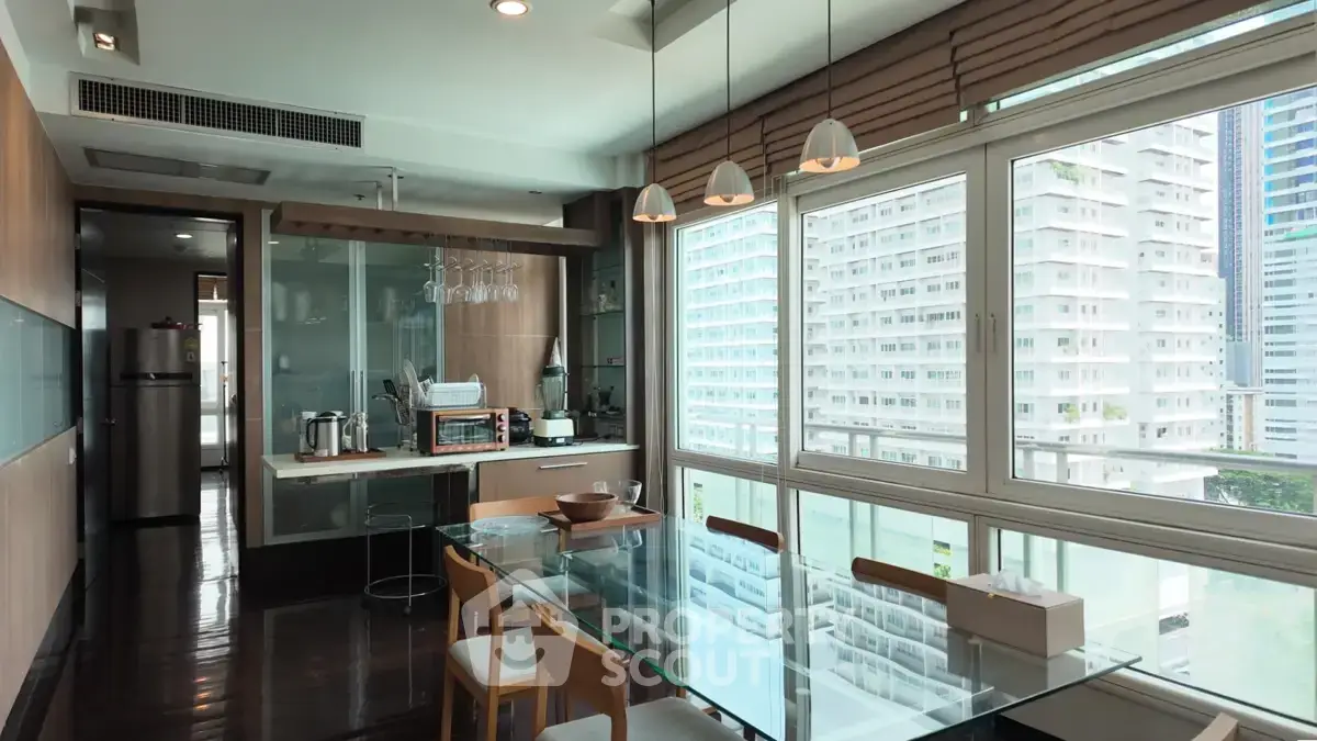 Modern dining area with glass table and city view, featuring open kitchen layout and stylish lighting.