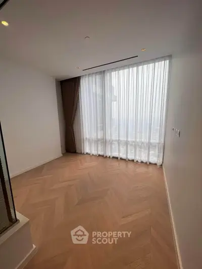Spacious empty room with elegant wooden flooring and large window with sheer curtains.
