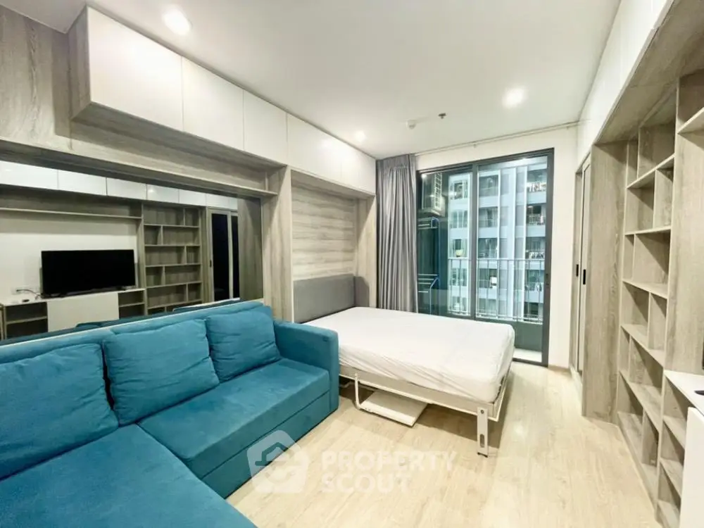 Modern studio apartment with blue sofa and bed, featuring large windows and built-in storage.