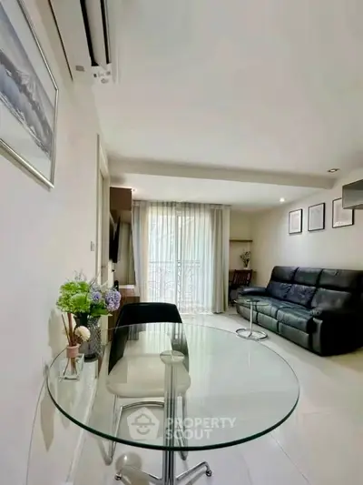Modern living room with glass table and black leather sofa, featuring elegant decor and natural light.
