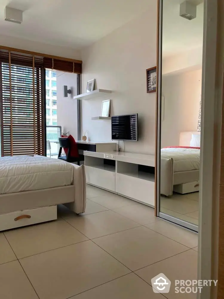 Spacious bedroom with large mirror wardrobe doors reflecting a well-lit room, modern furniture, and a glimpse of the city view through wooden blinds.