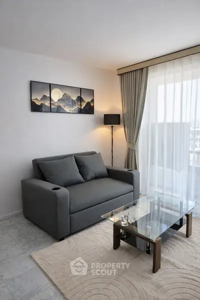 Modern living room with sleek gray sofa and glass coffee table, featuring mountain artwork and floor-to-ceiling windows.