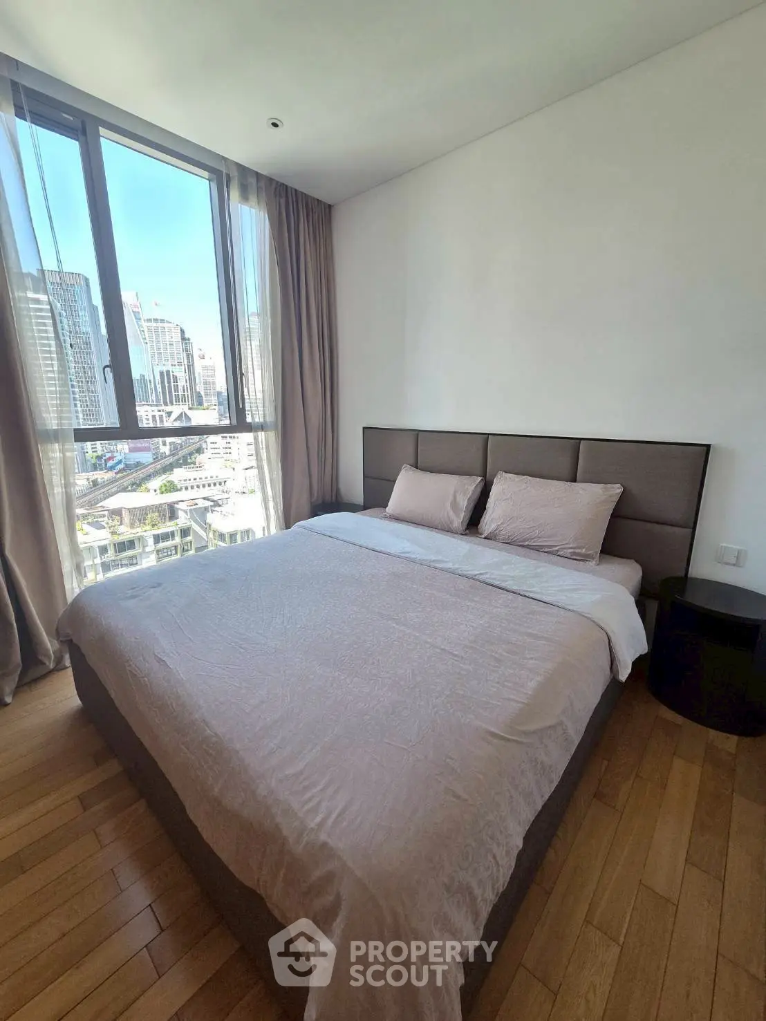 Modern bedroom with city view, featuring a large bed and wooden flooring in a high-rise apartment.