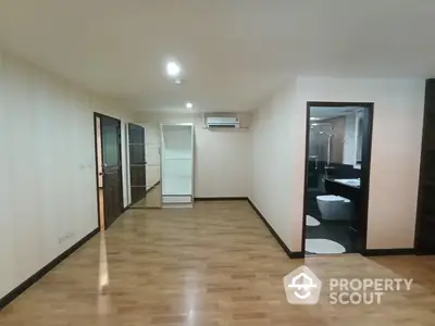 Spacious and well-lit apartment interior with gleaming hardwood floors, featuring an open layout and modern appliances, including a refrigerator and air conditioning unit.