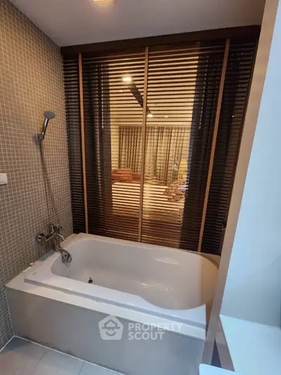 Elegant bathroom with a large bathtub, modern fixtures, and mosaic tile walls, complemented by warm wooden blinds and ambient lighting.