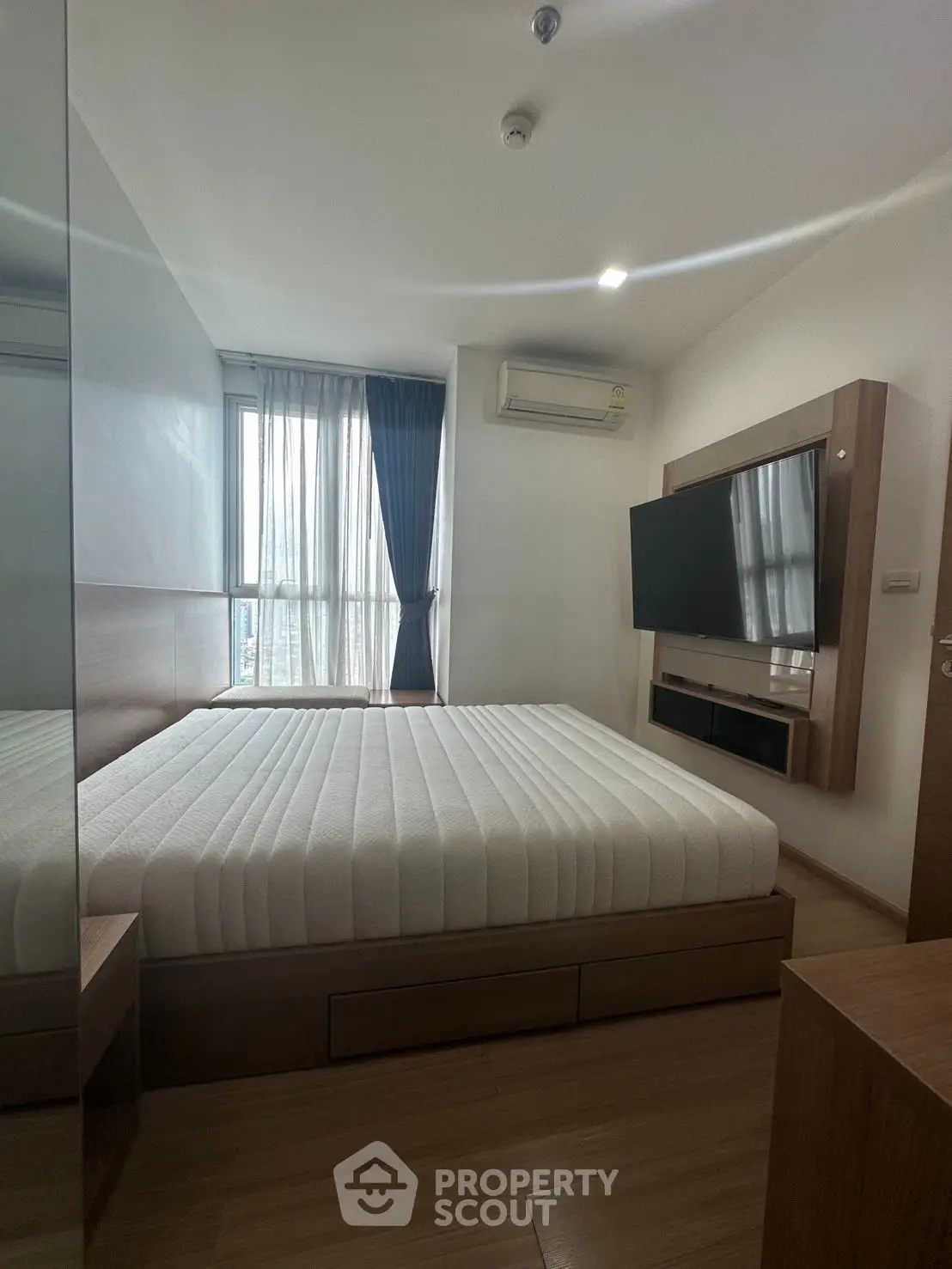 Modern bedroom with large window and wall-mounted TV, featuring sleek wooden furniture.