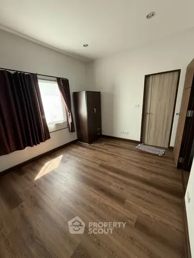 Spacious bedroom with wooden flooring and natural light, featuring a wardrobe and elegant curtains.