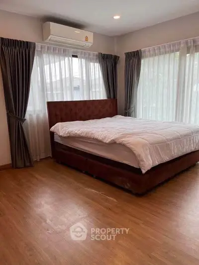 Spacious bedroom with large windows and elegant curtains, featuring a comfortable bed and wooden flooring.