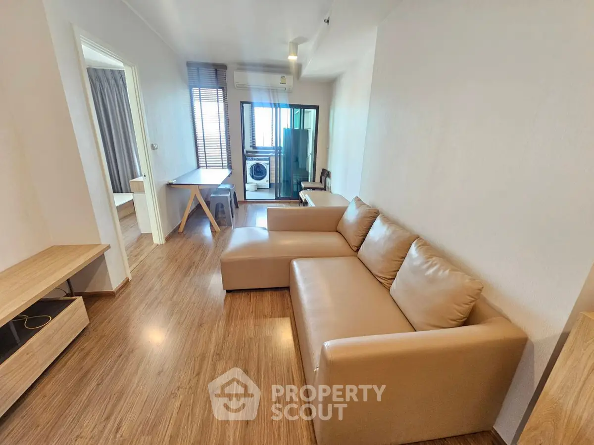 Modern living room with brown leather sofa and wooden flooring, featuring a view into a bedroom and laundry area.