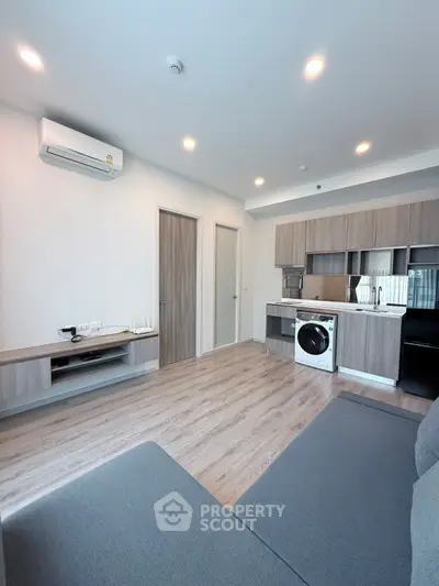 Modern living room with sleek kitchen and washing machine, featuring wooden flooring and ample lighting.