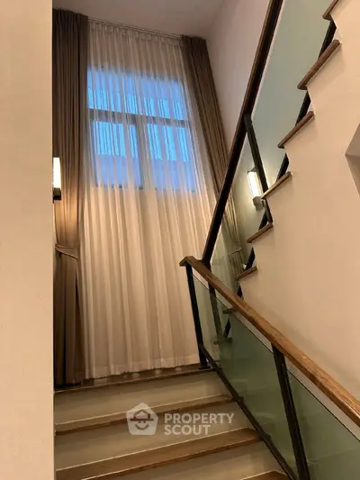 Elegant staircase with modern glass railing and tall window with curtains