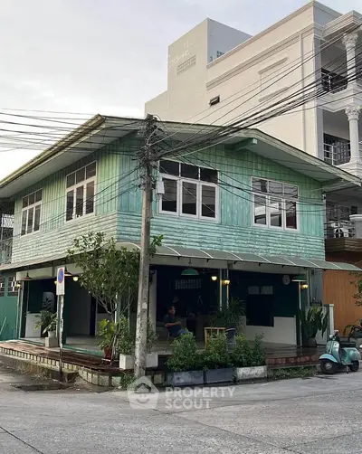 Charming vintage two-story house with green wooden exterior and lush plants on a quiet street corner.