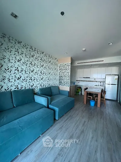 Modern living room with stylish wallpaper and teal sofas, open layout to kitchen area with dining table.