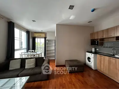 Spacious living room with modern kitchen and washing machine, featuring large windows and wooden flooring.