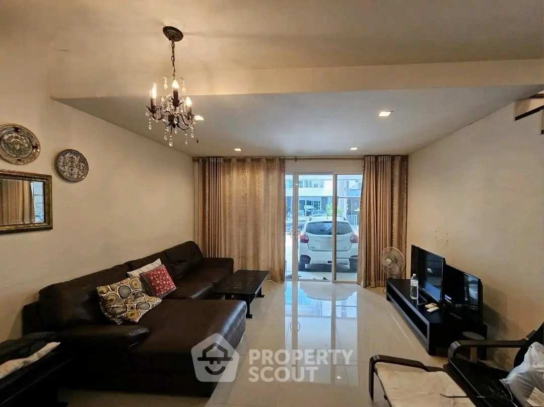 Spacious living room with elegant chandelier and modern furniture, featuring a large window with a view of parked cars.