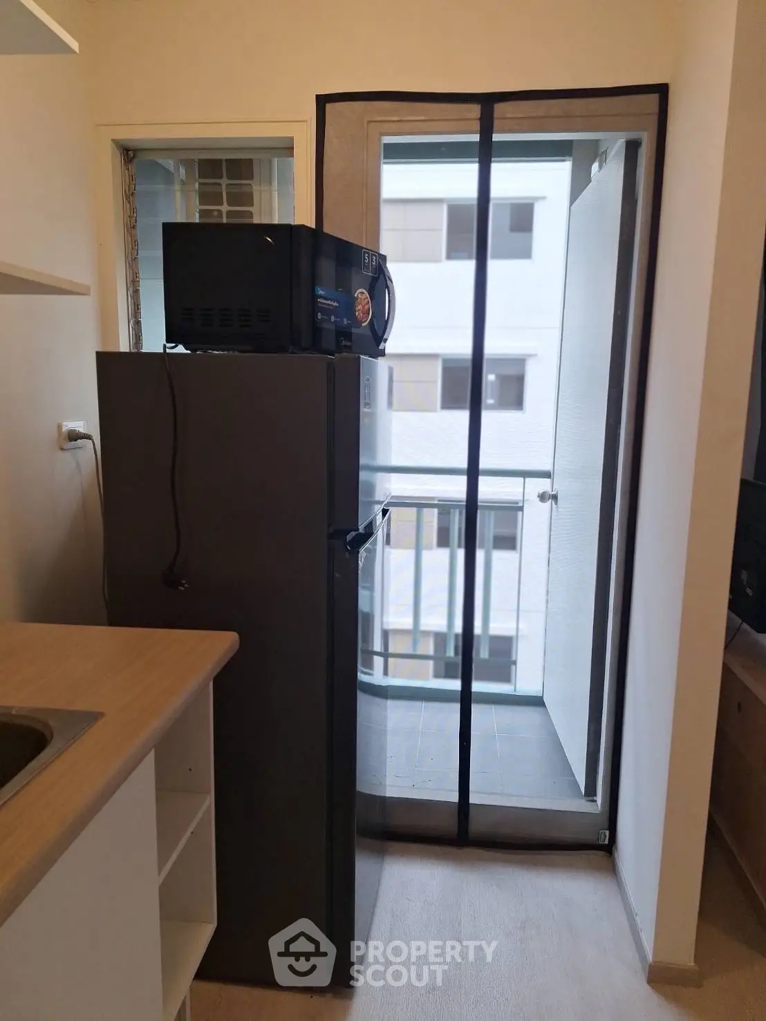 Compact kitchen with fridge and microwave leading to a small balcony with urban view.