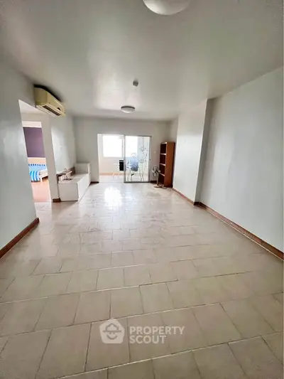 Spacious living room with tiled flooring and natural light, leading to a cozy bedroom.