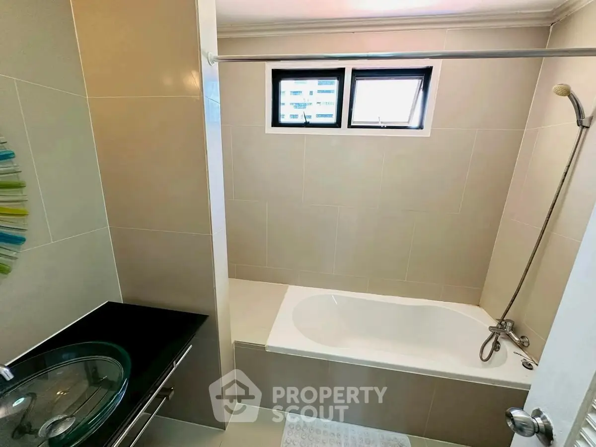 Modern bathroom with sleek design and built-in bathtub, featuring a stylish glass sink and neutral tiles.