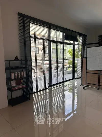 Modern living room with large windows and sleek blinds, featuring a stylish bookshelf and whiteboard.