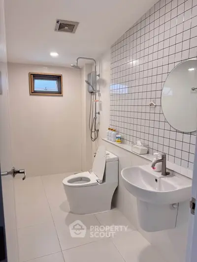 Modern bathroom with sleek fixtures and tiled walls, featuring a round mirror and compact shower area.