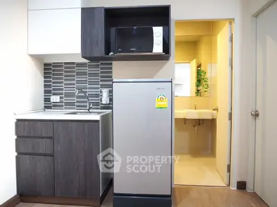 Modern compact kitchen with microwave and fridge adjacent to a bathroom, featuring sleek cabinetry and tiled backsplash.