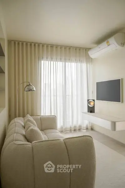 Modern living room with cozy beige sofa, wall-mounted TV, and floor lamp, featuring large window with sheer curtains.